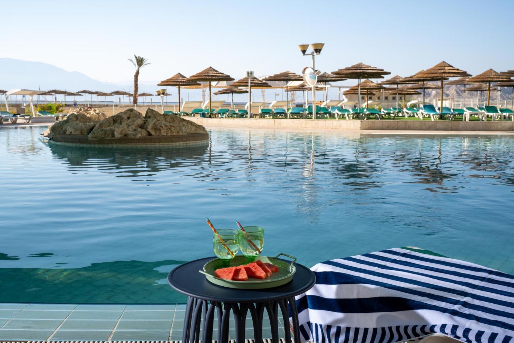 Leonardo Club Hotel Dead Sea Neve Zohar Exterior photo The photo depicts a tranquil swimming pool area at a resort or hotel. In the foreground, there is a small round table with a plate containing slices of watermelon and two green drinks. The background features a calm pool with a small rock island in t